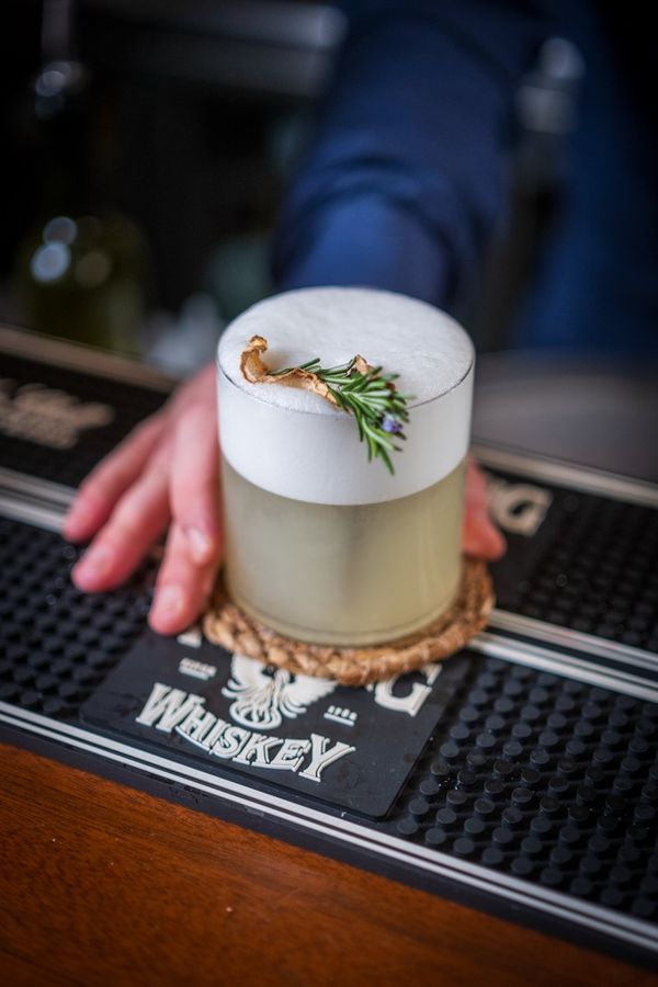 Foamy cocktail with a rosemary sprig set down on the bar