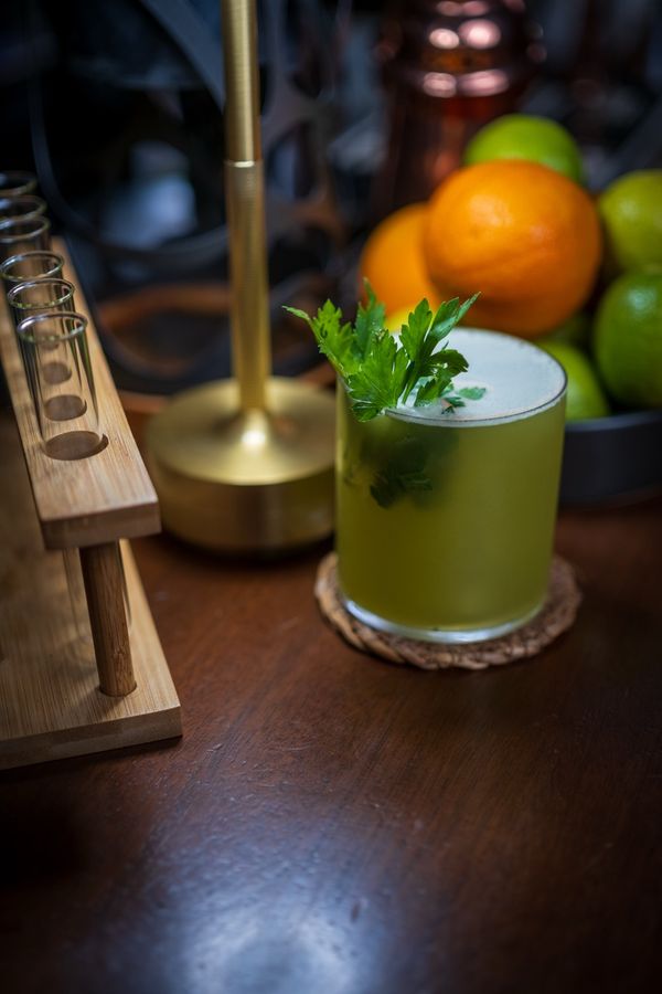 Green cocktail with a herb garnish, oranges and limes behind